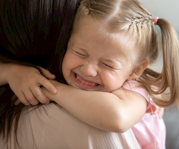 A young happy, smiling girl shown over the shoulder of a dark haired woman, hugging her tight