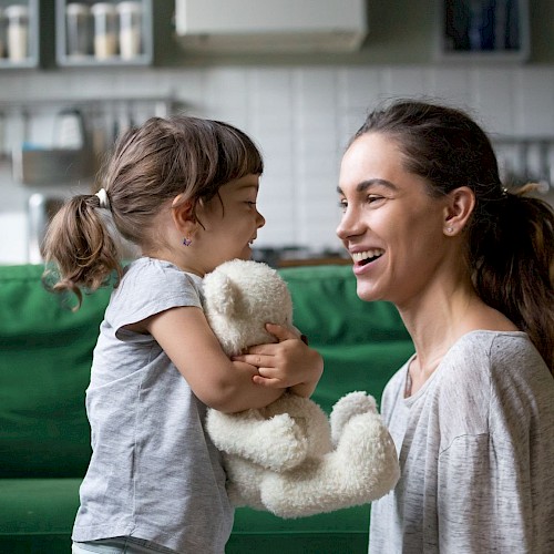 Happy and laughing young girl cuddling a teddy with a smiling young woman next to her