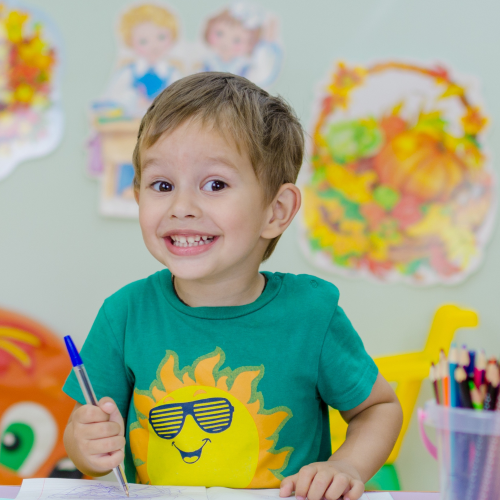 Cheeky boy holding a blue pen in front of a blurred backdrop of children's artwork