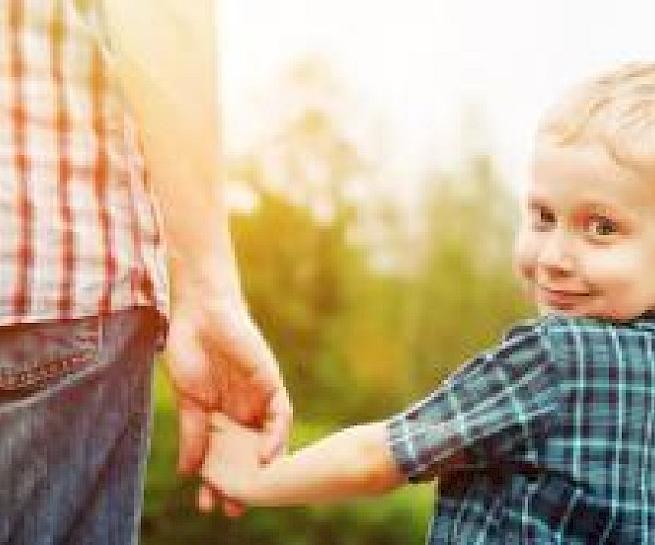 young boy peering over his should whilst holding hands with a man in a red checked shirt