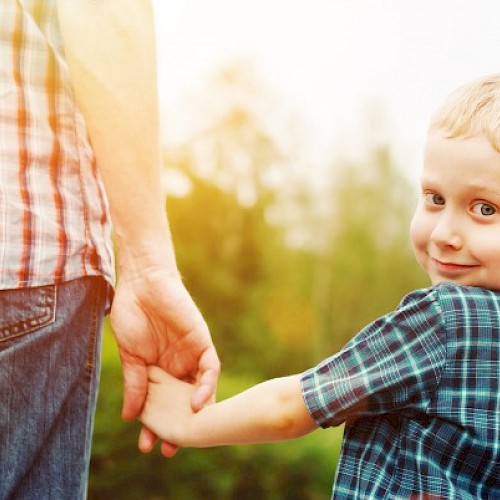 young boy peering over his should whilst holding hands with a man in a red checked shirt