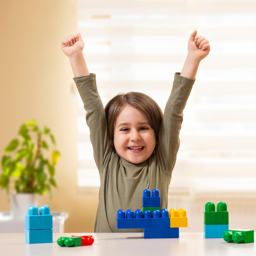 Happy young girl with arms in the air infront of some building bricks on a table